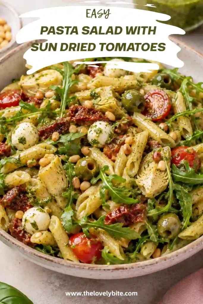 Mediterranean pasta salad with penne, sun-dried tomatoes, artichokes, mozzarella balls, olives, cherry tomatoes, and homemade arugula pesto in a large serving bowl.